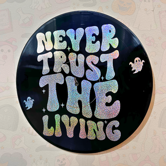 A circular black vinyl record or plaque featuring the phrase “NEVER TRUST THE LIVING” in large holographic glitter letters, decorated with small ghost and star graphics, shown against a patterned beige background.