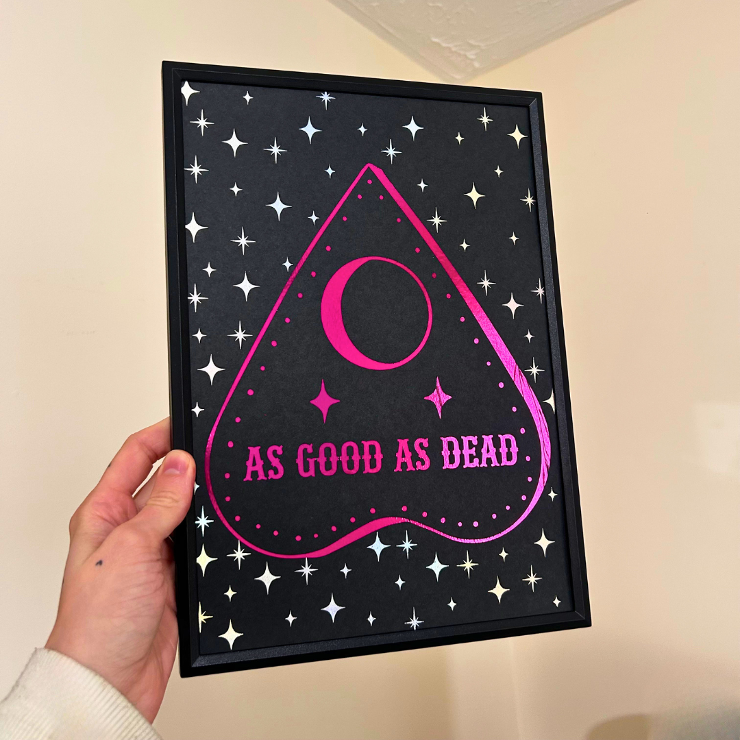 A hand holds a framed gothic-style art print featuring a black background with white stars, a pink heart-shaped outline, a pink crescent moon, and the text 'AS GOOD AS DEAD' in pink. The frame is black, and the background wall is a warm beige.