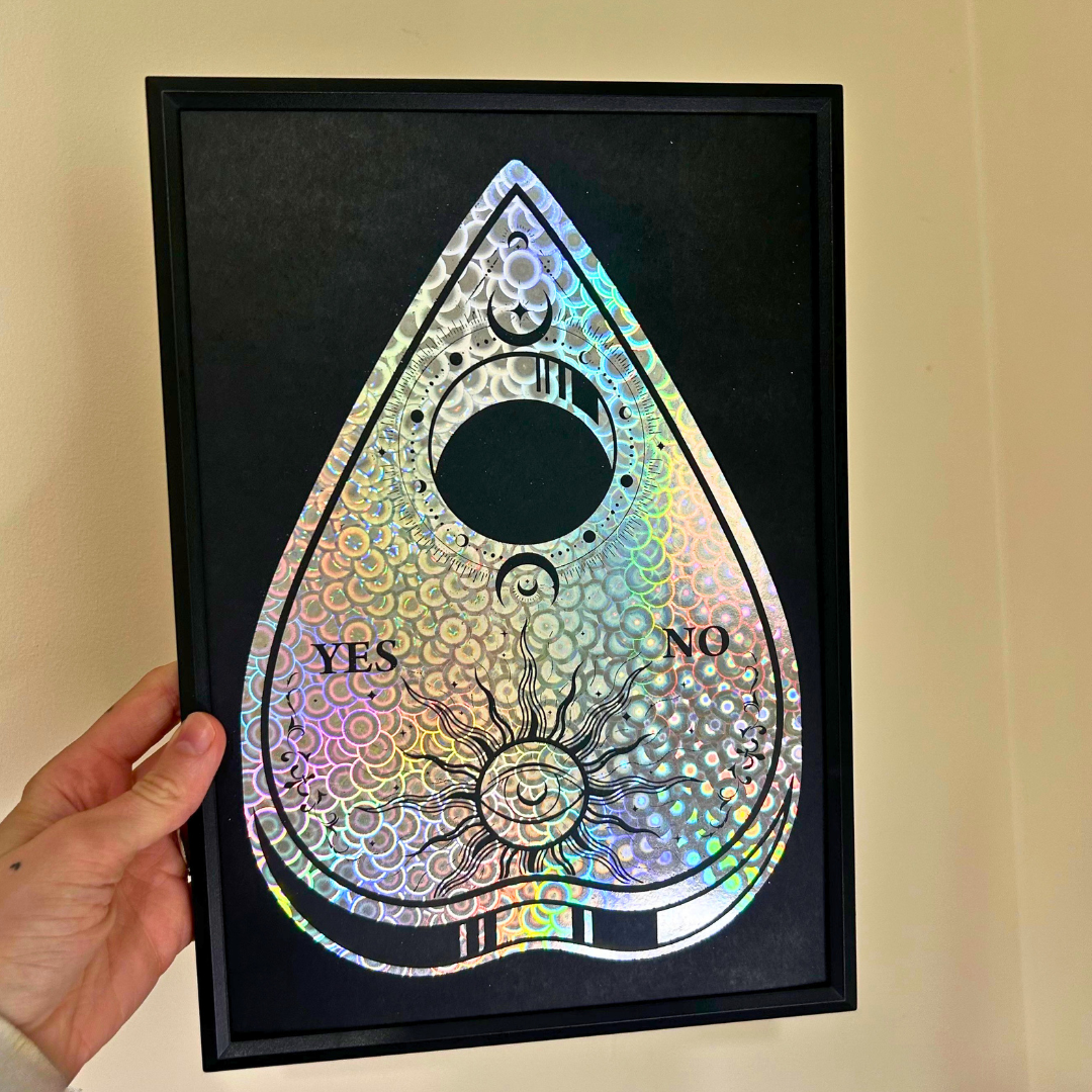 A black-framed teardrop-shaped board with an iridescent holographic pattern, featuring a central circular cutout, the words YES and NO, and a sun-and-moon motif. A hand is holding the frame against a beige wall.