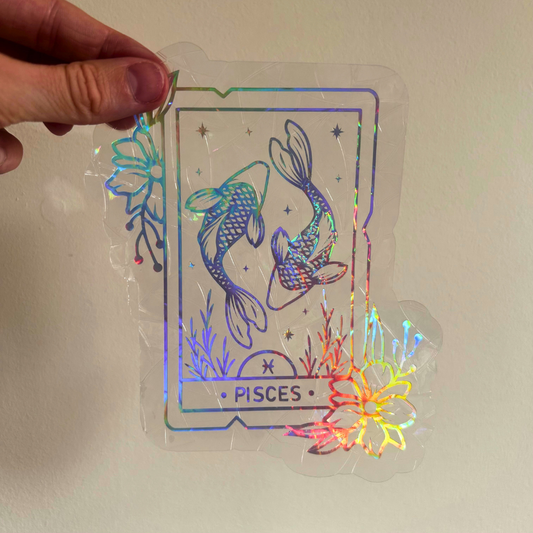 A hand holds a clear holographic sticker or decal shaped like a Pisces tarot card, featuring two koi fish and iridescent rainbow reflections, against a plain beige wall.