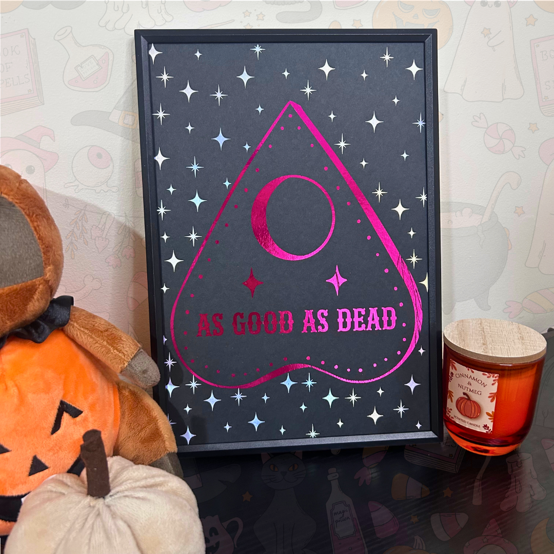 A framed gothic-style art print featuring a pink occult-style heart shape with a crescent moon and the text 'AS GOOD AS DEAD' on a dark gray starry background. The frame is displayed on a reflective surface next to a plush toy, a small pumpkin, and an amber candle.