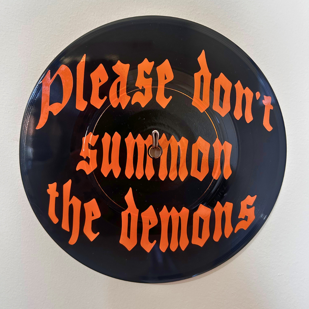 A circular black vinyl record wall hangingwith the phrase 'Please don't summon the demons' written in orange Gothic lettering