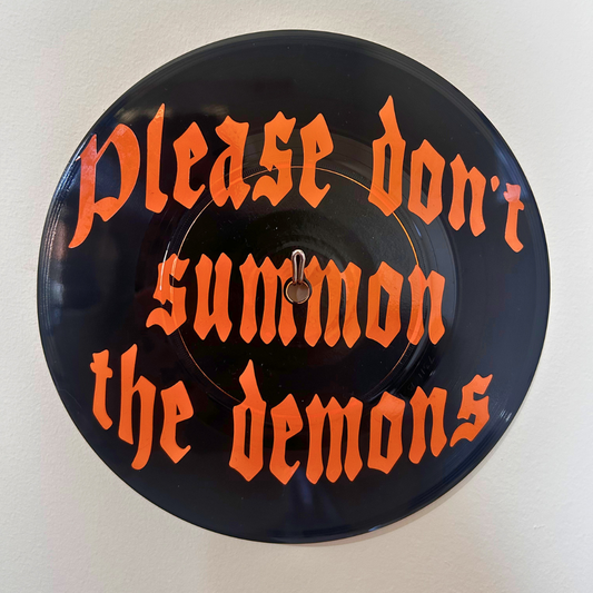 A circular black vinyl record wall hangingwith the phrase 'Please don't summon the demons' written in orange Gothic lettering