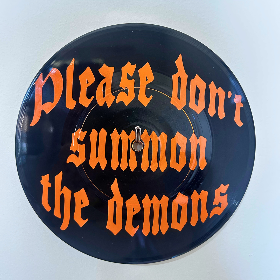 A circular black vinyl record wall hanging featuring the phrase 'Please don't summon the demons' in a bright orange gothic font, with a small metal hand in the center, shown against a plain off-white background.