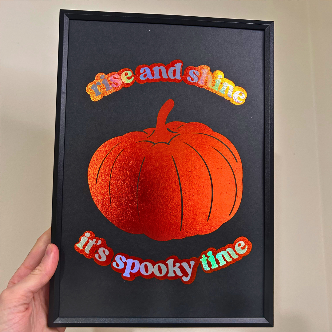 A hand holds a black-framed art print featuring a textured orange pumpkin and the phrase 'rise and shine it's spooky time' in colorful letters on a black background.