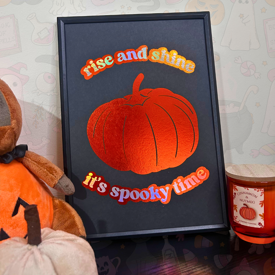 A framed decorative sign with a black background, a large orange pumpkin illustration, and the words “rise and shine” at the top and “it’s spooky time” at the bottom. The frame is styled on a tabletop with a plush pumpkin, a candle, and a patterned wall in the background.