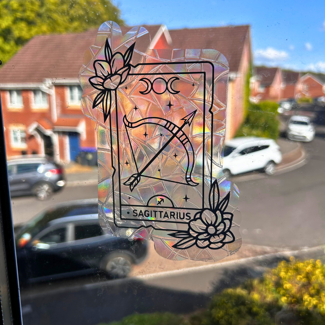 A holographic, iridescent vinyl decal featuring a black line-art Tarot card design with a bow and arrow, stars, floral corner accents, and the text 'SAGITTARIUS' at the bottom. The decal is shown applied to a window with a blurred street scene in the background.