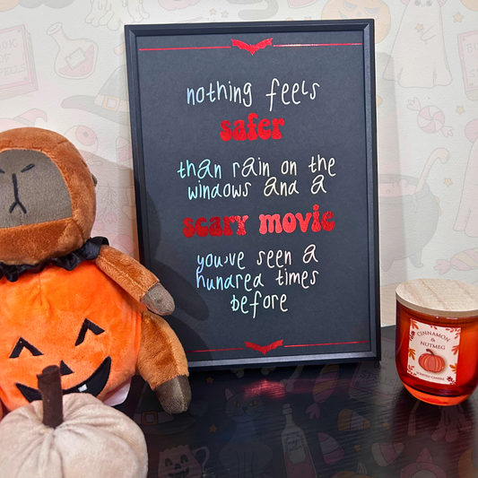 A framed black chalkboard sign with a Halloween-themed quote in white, red, and light blue lettering, styled on a dark tabletop next to a plush pumpkin and a candle.