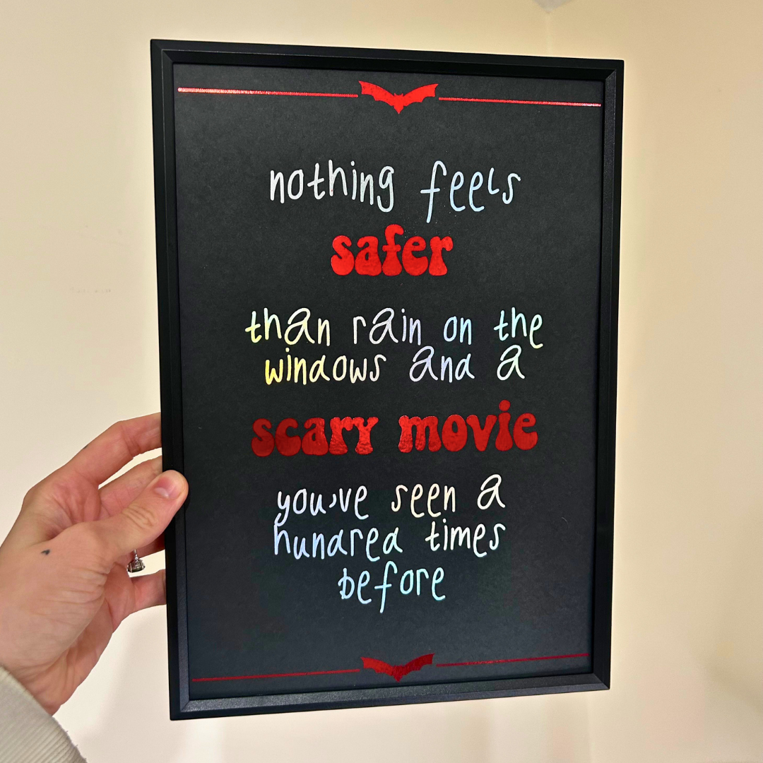 A hand holding a black framed chalkboard sign with a horror-themed quote about scary movies and rain, against a plain beige wall.