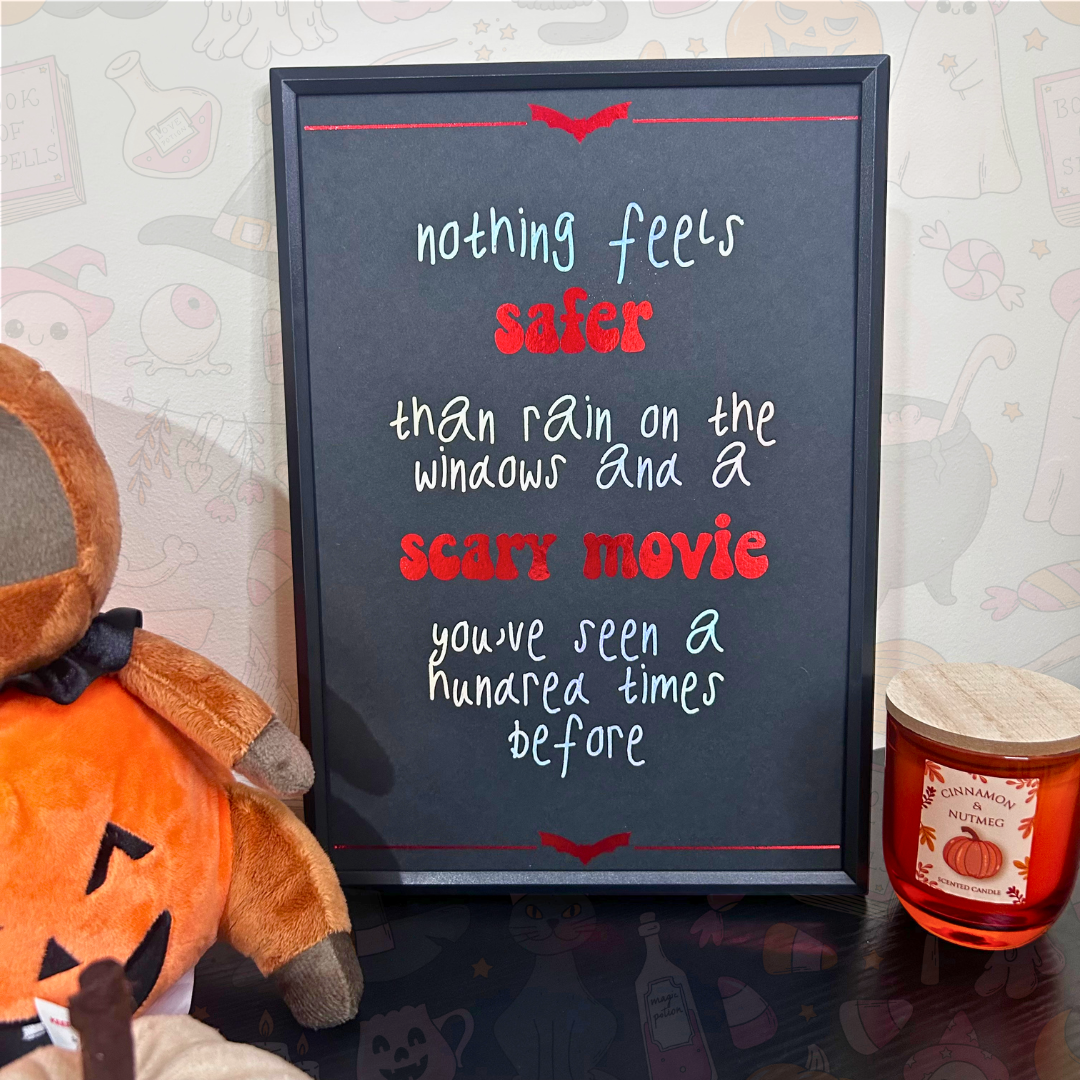 A framed black chalkboard sign with a Halloween quote in white and red lettering, styled on a tabletop with a plush pumpkin and a candle nearby against a patterned wallpaper background.