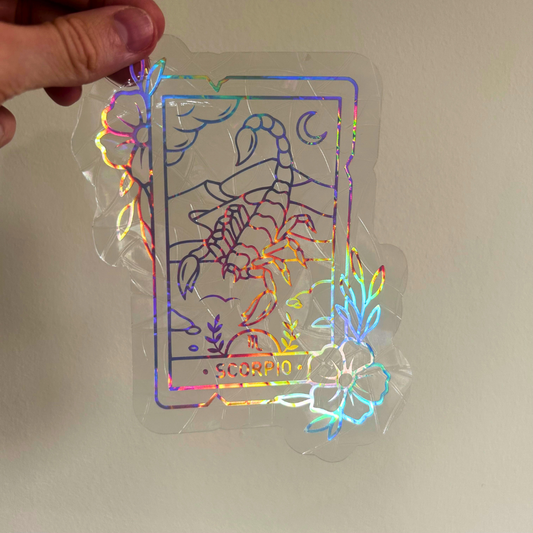 A hand holds a clear acrylic sticker featuring a silver line art tarot card design labeled 'SCORPIO' with holographic rainbow foil accents, against a plain off-white wall.