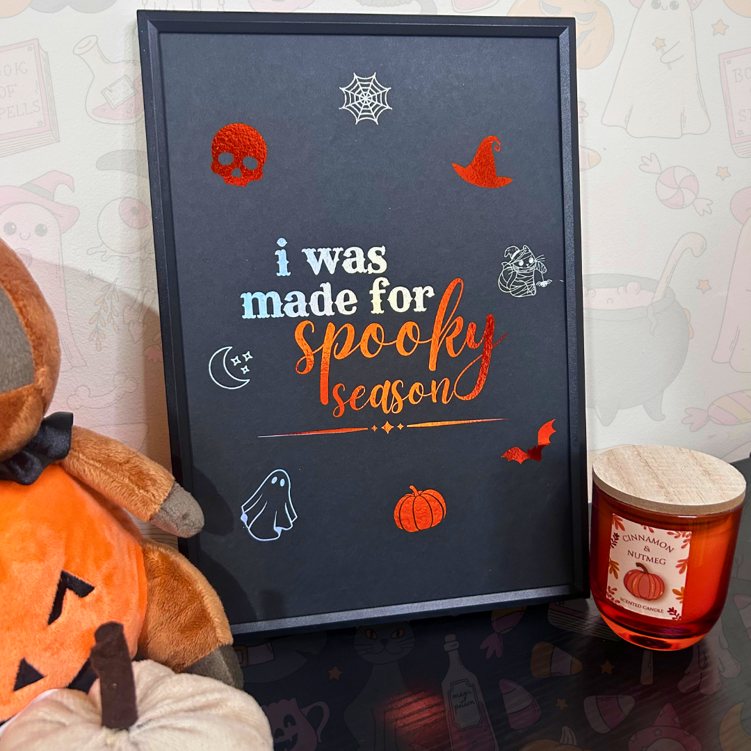 A framed Halloween-themed wall art print with the text 'i was made for spooky season' in white and orange lettering, surrounded by small spooky illustrations like a ghost, pumpkin, spiderweb, and witch hat. The frame is styled on a tabletop next to a plush jack-o'-lantern and a candle, against a patterned wall.