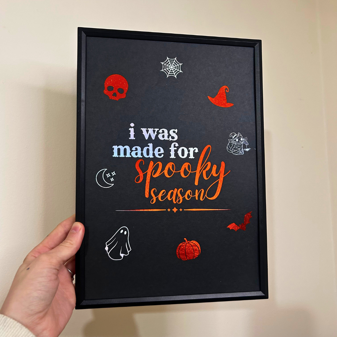 A hand holding a black-framed Halloween art print with the text 'i was made for spooky season' in white and orange, surrounded by small red and white spooky icons like a skull, witch hat, spiderweb, pumpkin, and ghosts, against a beige wall.