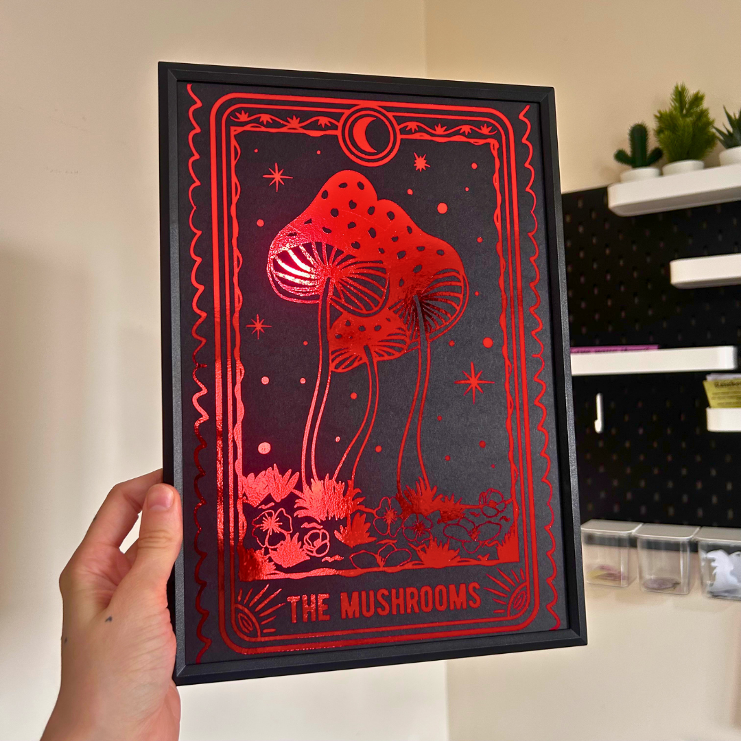 A hand holds a black-framed art print featuring red mushroom illustrations and the text 'THE MUSHROOMS' against a dark background, with a home interior visible behind it.