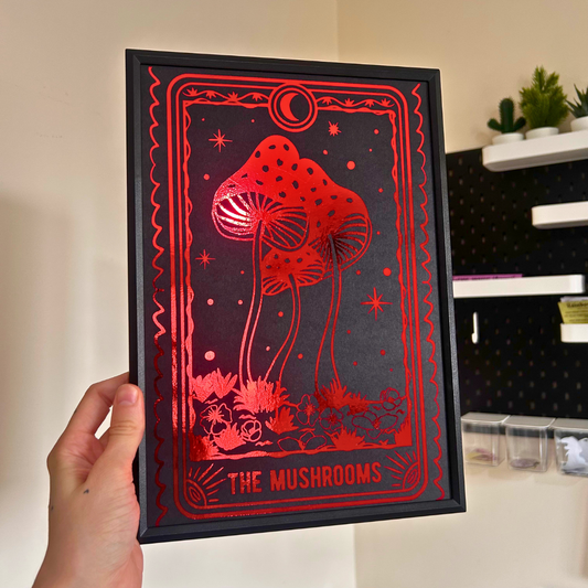 A hand holds a black-framed art print featuring red mushroom illustrations and the text 'THE MUSHROOMS' against a dark background, with a home interior visible behind it.