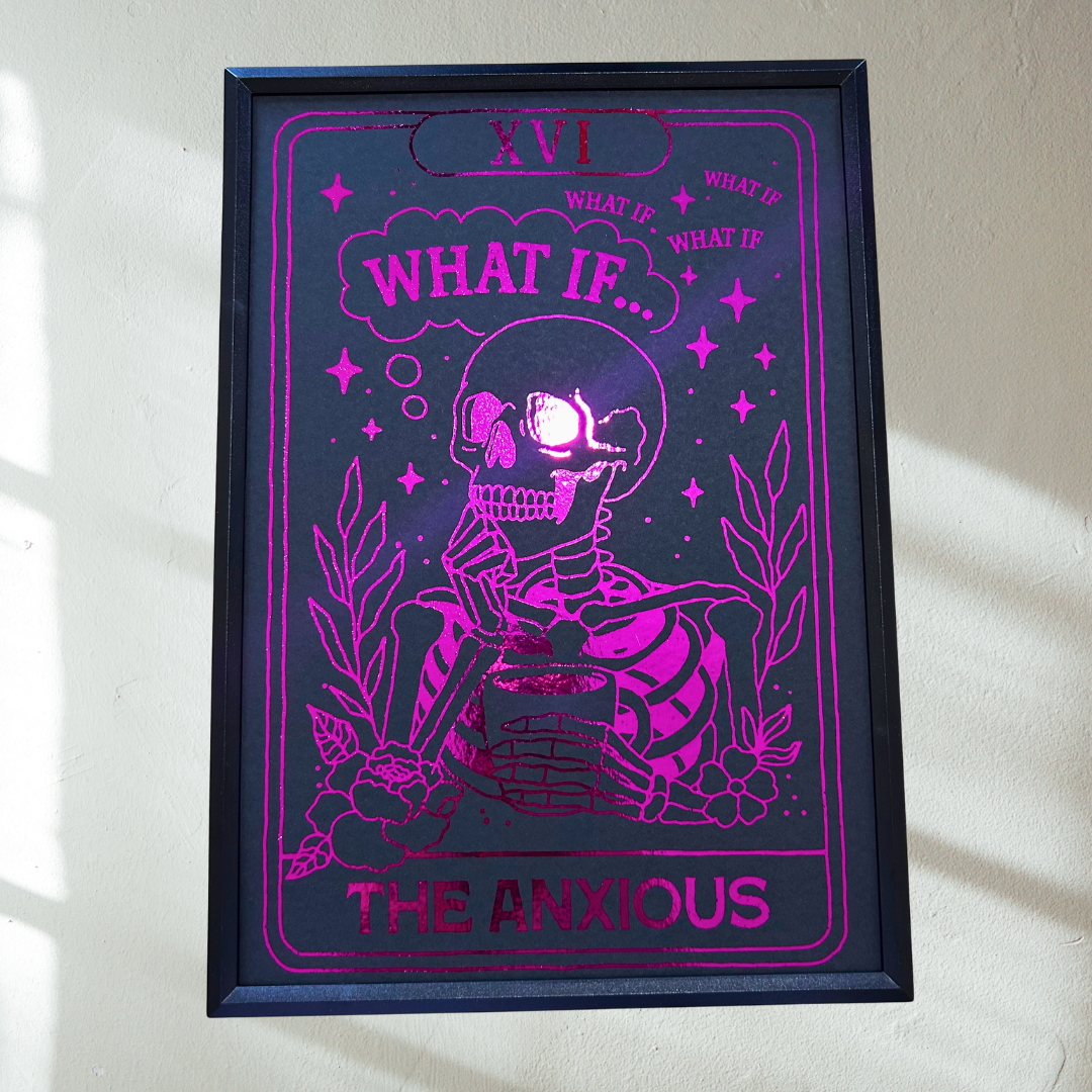 A framed art print featuring a pink and black illustration of a skeleton sitting in a chair with the text 'WHAT IF.