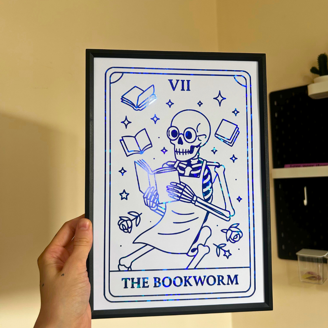 A hand holds a framed tarot-style art print featuring a blue skeleton bookworm illustration labeled 'VII THE BOOKWORM' on a white background, with a black frame against a beige indoor wall.