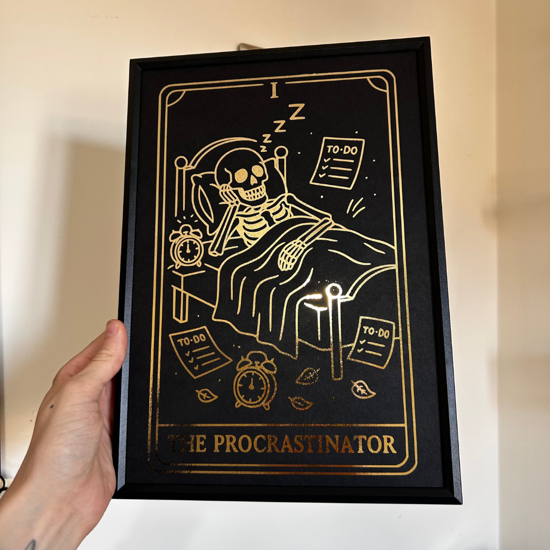 A hand holds a black-framed art print featuring a gold tarot-style illustration of a skeleton sleeping in bed with the title 'THE PROCRASTINATOR' at the bottom, against a plain beige wall.