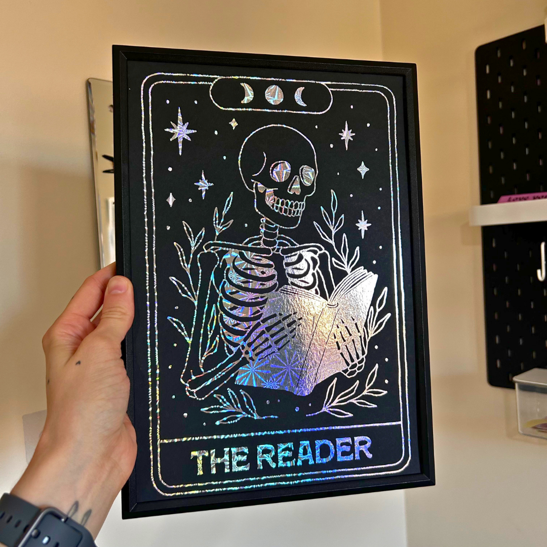 A hand holds a black tarot card art print featuring a skeleton reading a book, styled like 'The Reader' card with holographic foil text and moon phases at the top. The print is framed in black and set against a home interior background.