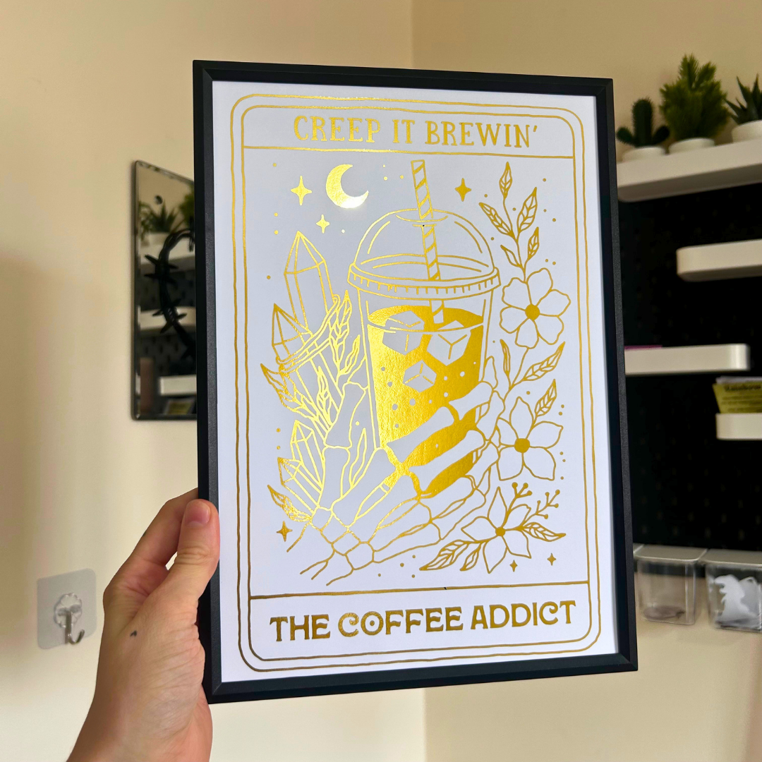 A hand holds a black-framed art print featuring a gold foil design of a coffee cup with the text 'CREEP IT BREWIN'' at the top and 'THE COFFEE ADDICT' at the bottom, against a beige wall background.