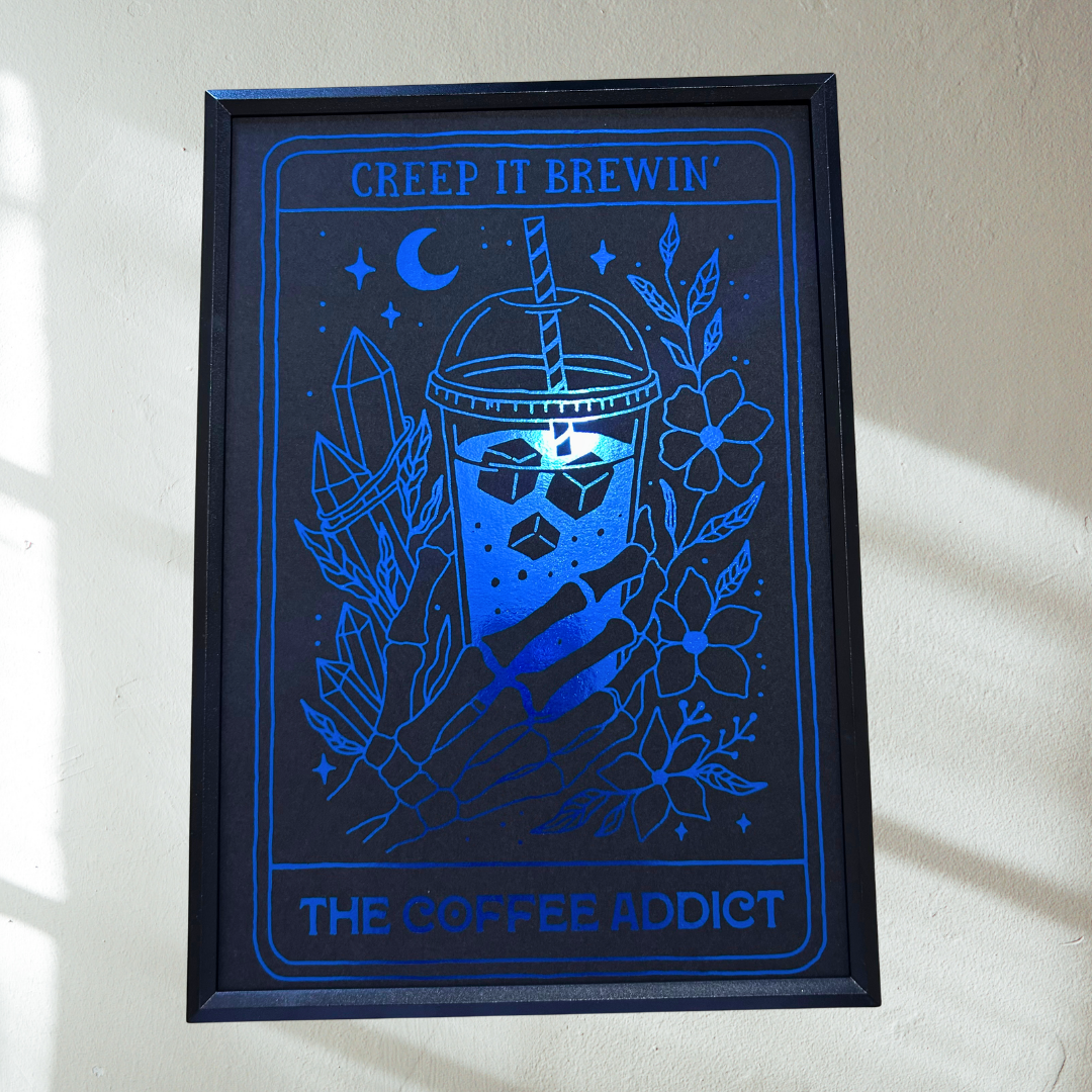 A framed art print featuring a stylized blue coffee cup with a skull graphic, surrounded by crystals, flowers, and stars, with the text 'CREEP IT BREWIN' at the top and 'THE COFFEE ADDICT' at the bottom, displayed on a light wall.