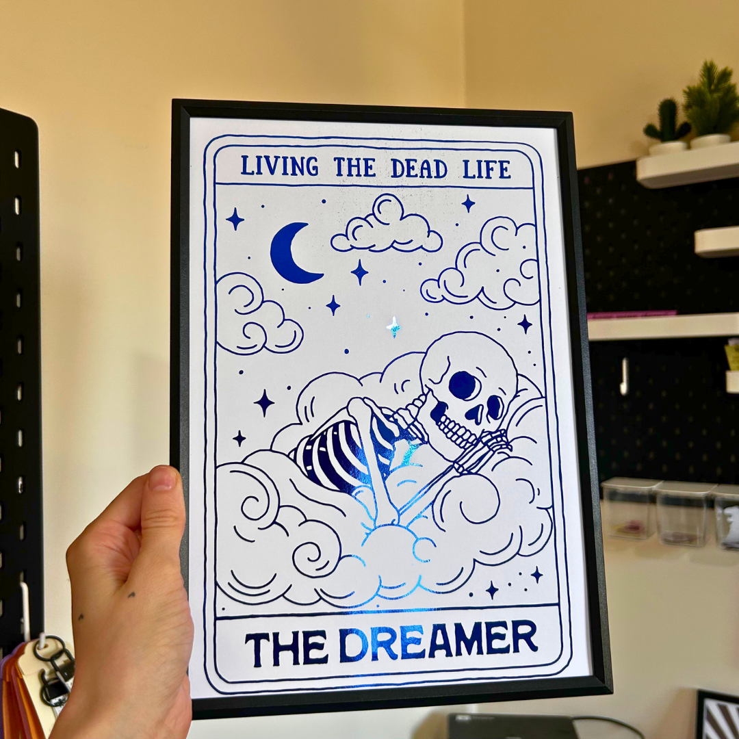 A hand holds a framed art print featuring a blue line-art illustration of a skeleton resting on clouds, with a crescent moon and stars. The text reads 'LIVING THE DEAD LIFE' at the top and 'THE DREAMER' at the bottom.
