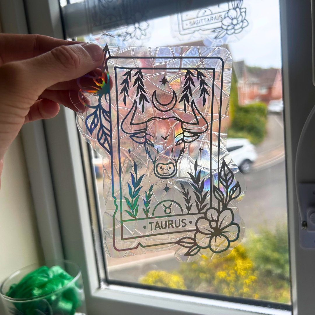 A hand holds a clear holographic Taurus tarot card sticker with silver line art in front of a window, showing its iridescent reflective properties against an outdoor street view.