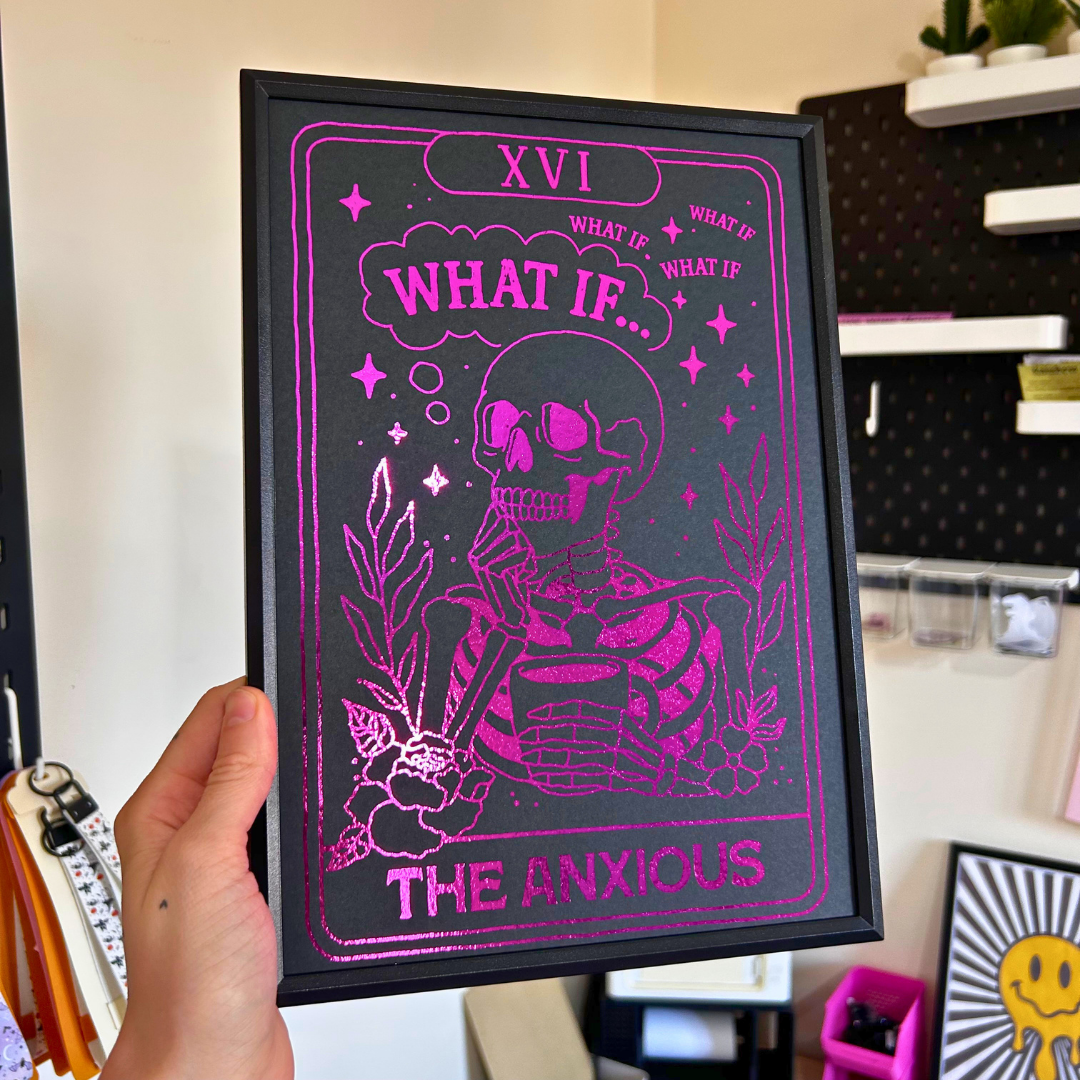 A hand holds a black-framed art print featuring a pink and black illustration of a skeleton with the text 'XVI WHAT IF.