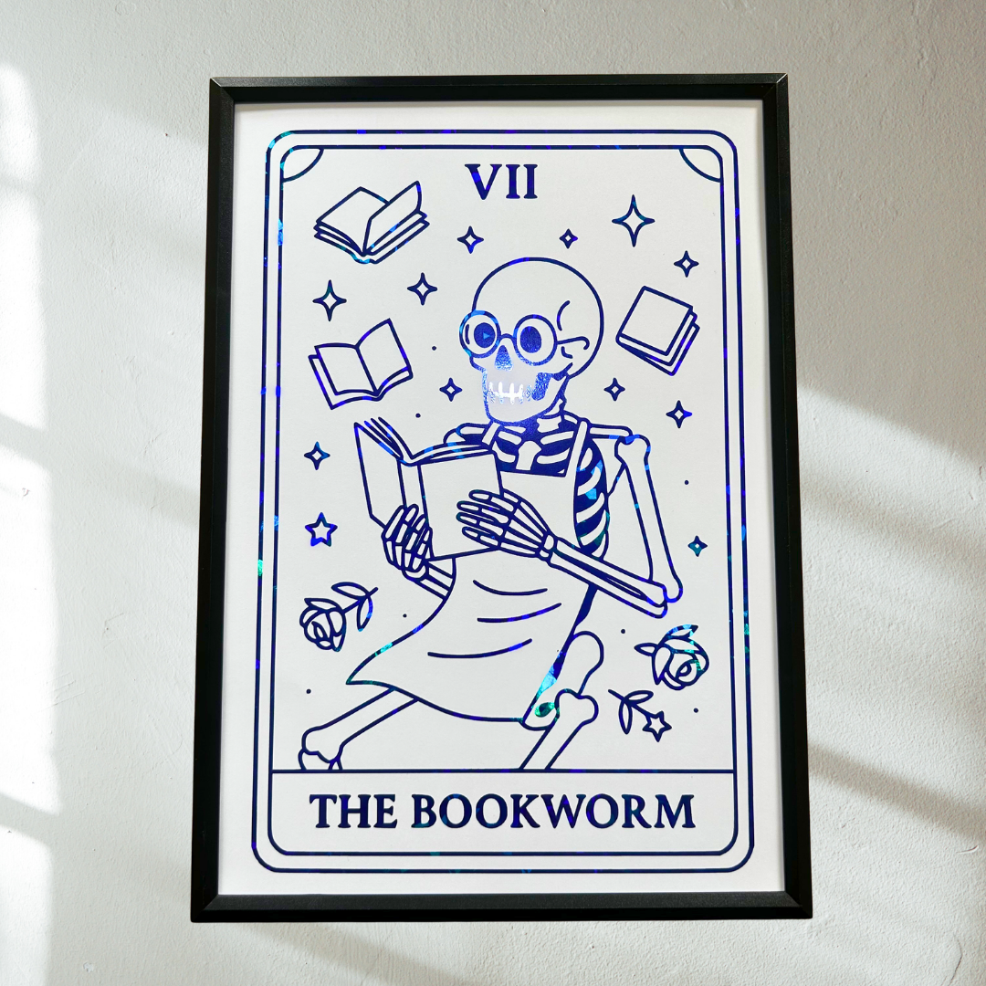 A framed tarot-style art print featuring a blue line-drawn skeleton bookworm with the Roman numeral VII and the text 'THE BOOKWORM' on a white background, hung on a light wall with soft window shadows.