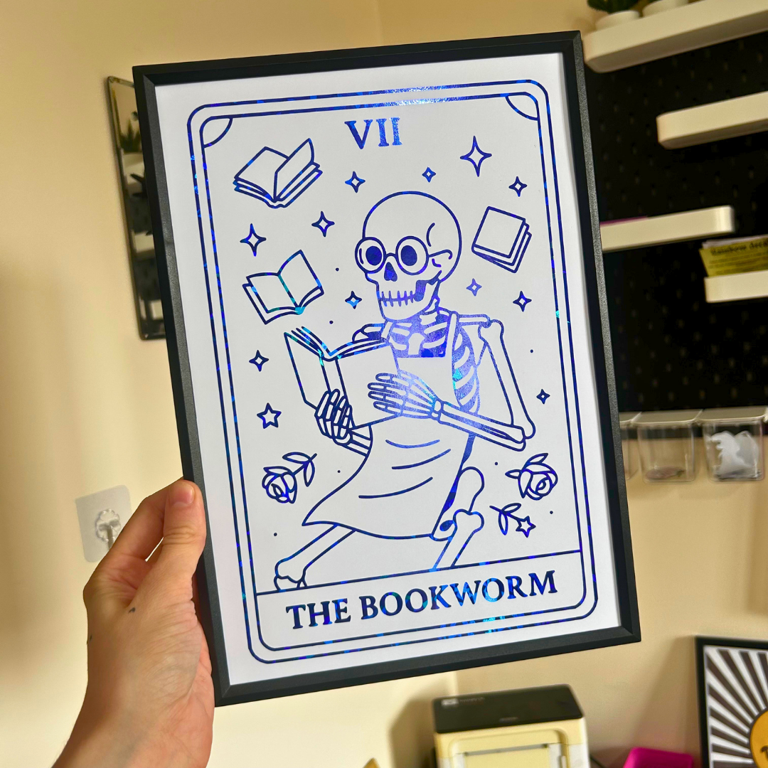 A hand holds a black-framed art print featuring a blue line-art illustration of a skeleton bookworm surrounded by books and roses, with the Roman numeral VII and the text 'THE BOOKWORM' on a white background.