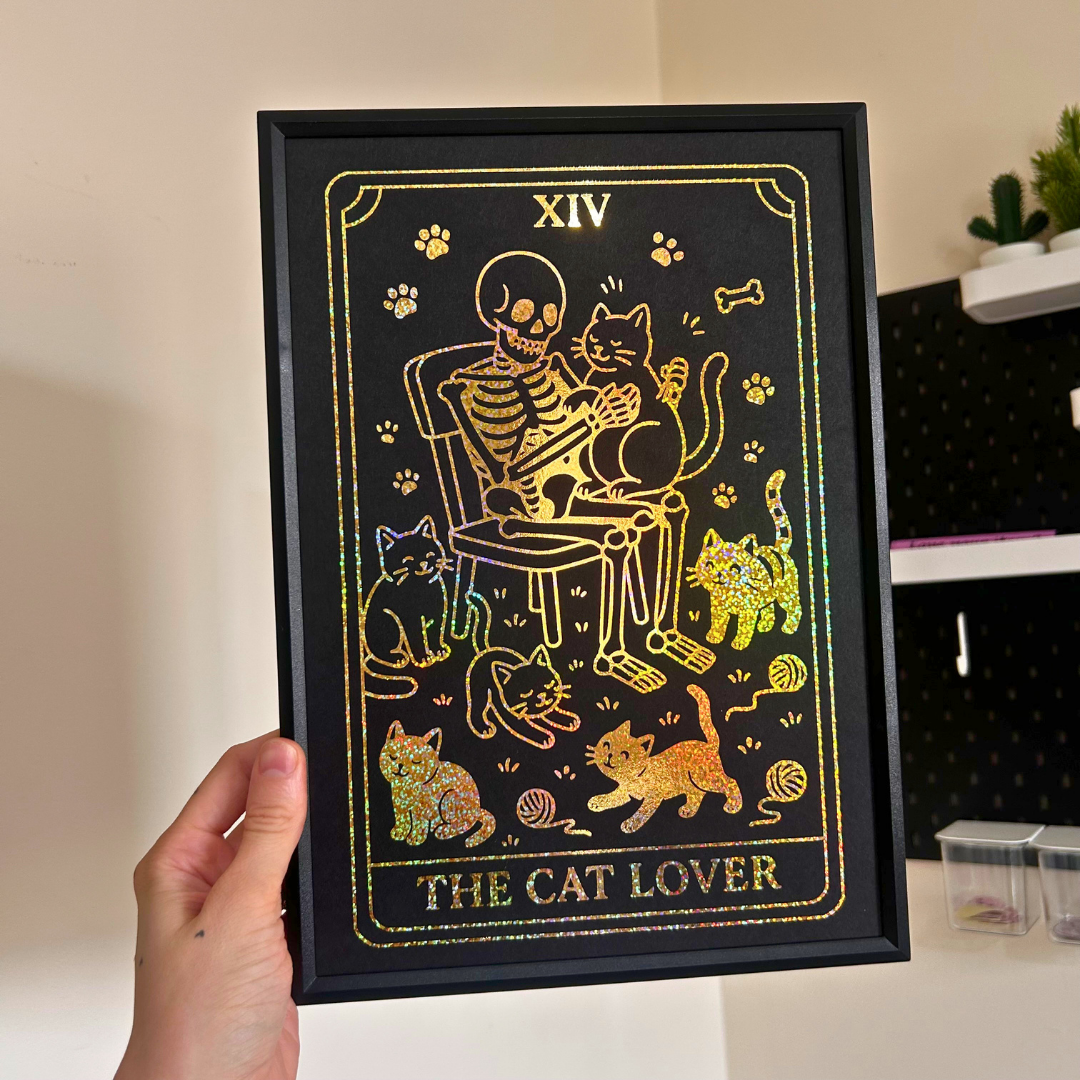 A hand holds a black-framed art print featuring a tarot card design with the text 'XIV' at the top and 'THE CAT LOVER' at the bottom. The artwork shows a skeleton sitting on a chair with several cats, rendered in gold and iridescent glitter on a black background.