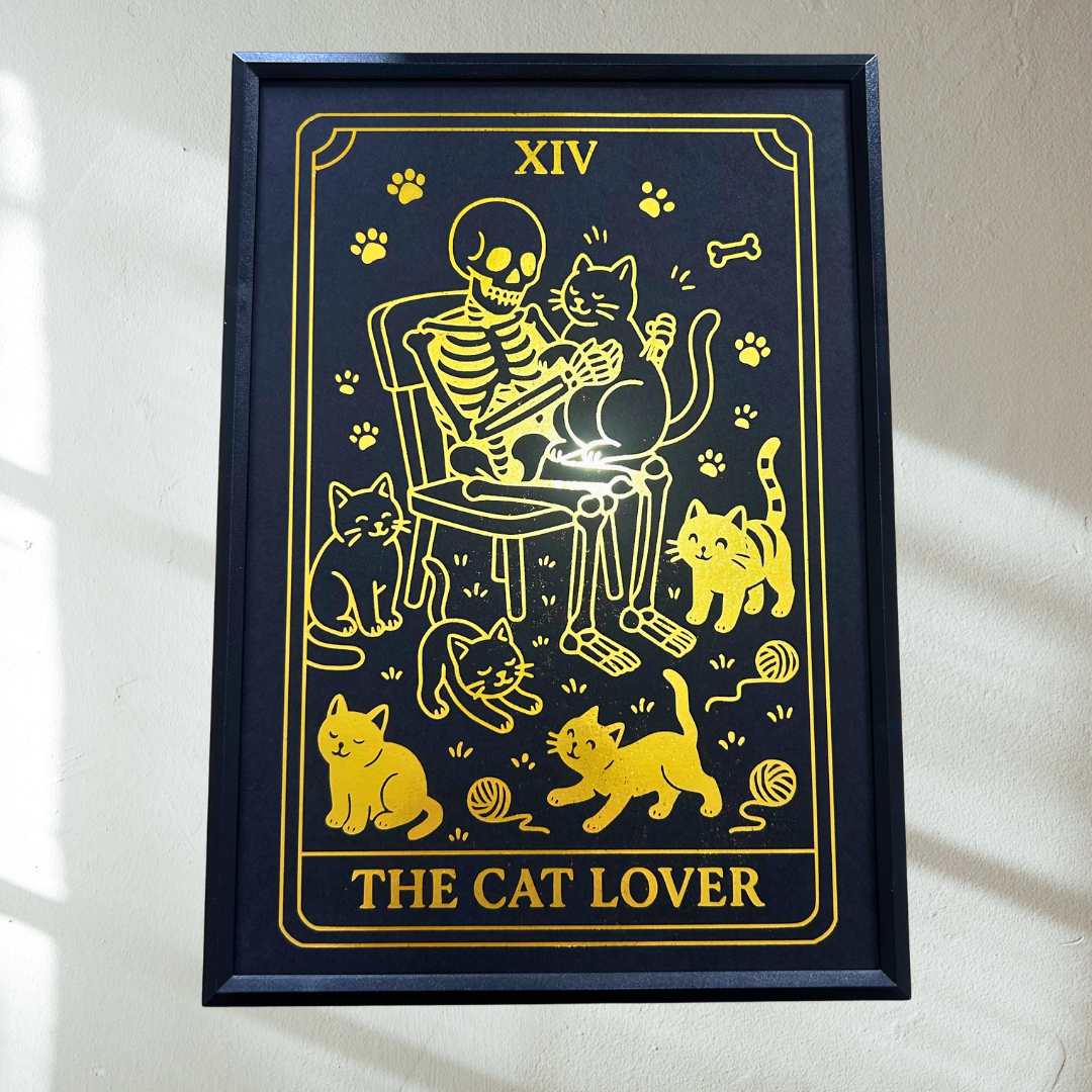 A framed art print featuring a tarot card design titled 'THE CAT LOVER' with a black background and yellow illustration of a skeleton sitting on a chair with multiple cats around it. The frame is mounted on a light-colored wall with soft sunlight shadows.