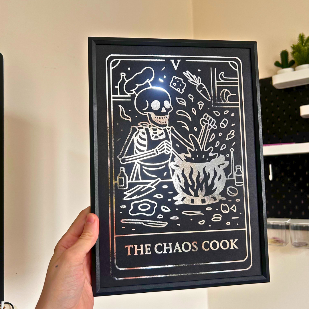 A hand holds a black-framed art print featuring a stylized skeleton chef cooking at a stove, with the text 'THE CHAOS COOK' at the bottom. The print has a dark background with white line art and text, shown against an indoor wall background.