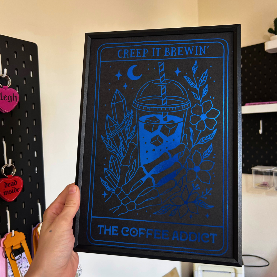 A hand holds a black-framed art print featuring a blue line art design of a coffee cup with crystals, flowers, and a crescent moon, with the text 'CREEP IT BREWIN'' at the top and 'THE COFFEE ADDICT' at the bottom.