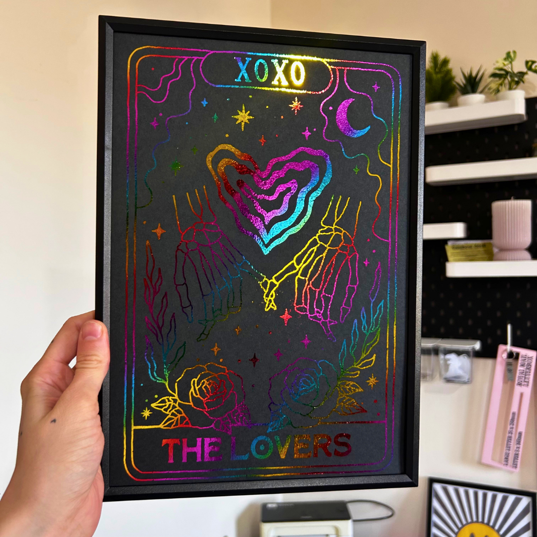 A hand holds a black-framed tarot card art print featuring 'The Lovers' design with holographic rainbow glitter lines on a black background. The scene is set in a home interior with shelves and decor visible in the background.