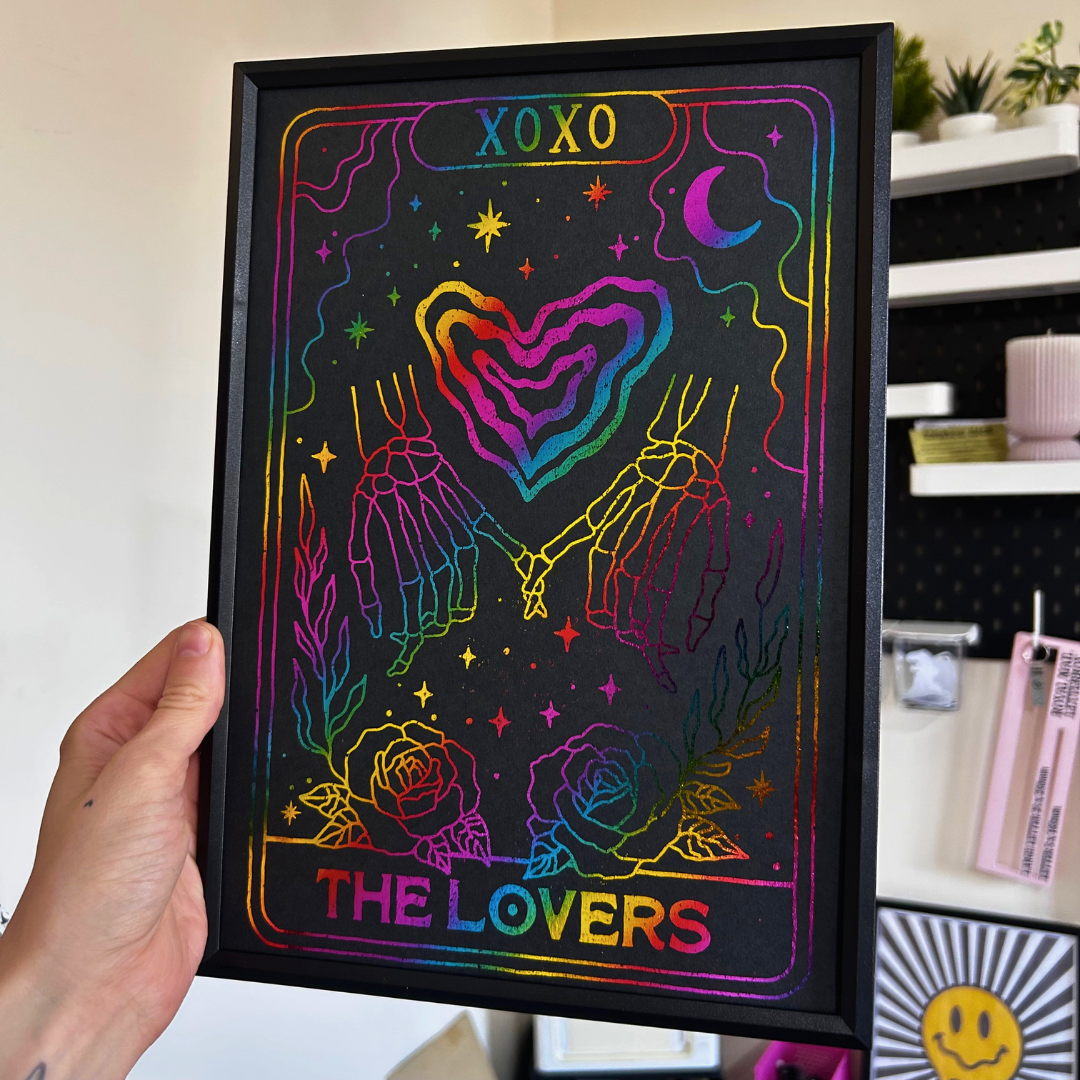 A hand holds a black tarot card art print styled as 'The Lovers' with a rainbow holographic foil effect. The print features skeletal hands holding, a heart, stars, and a crescent moon on a black background, framed in black.