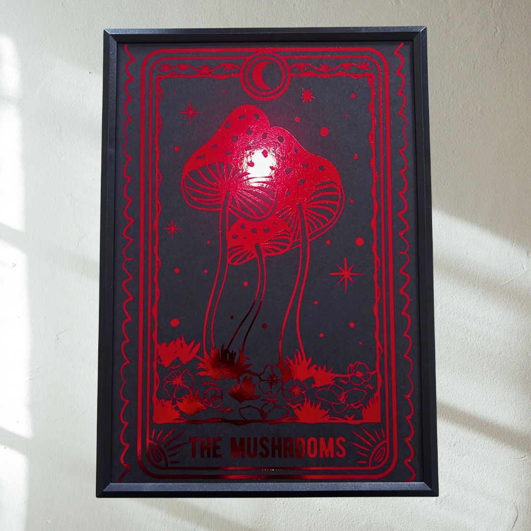 A framed art print featuring a red mushroom illustration on a dark background, hung on a light-colored wall with soft window shadows.