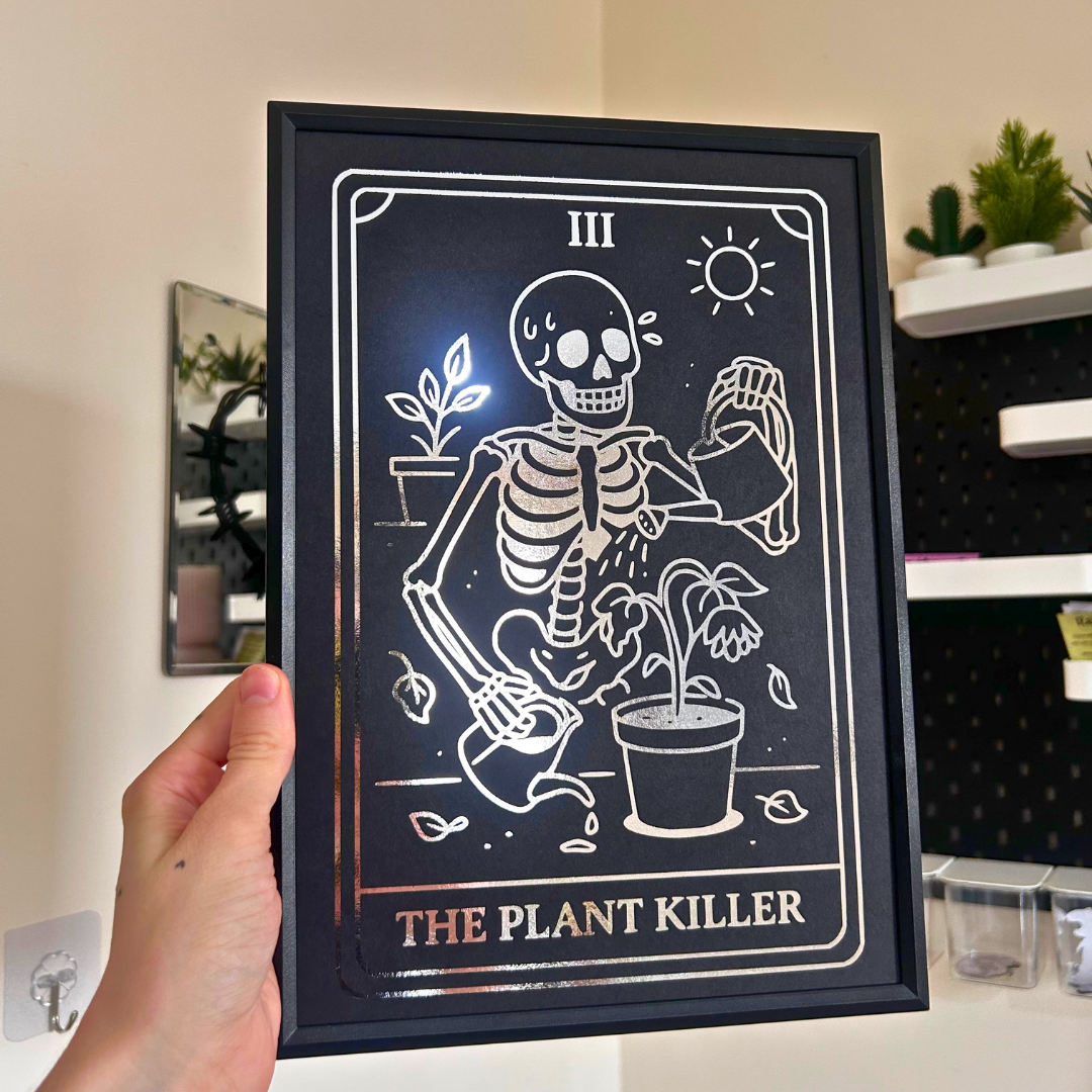 A hand holds a black framed art print featuring a white tarot-style skeleton illustration watering a plant, with the text 'THE PLANT KILLER' and 'III' on a dark background.