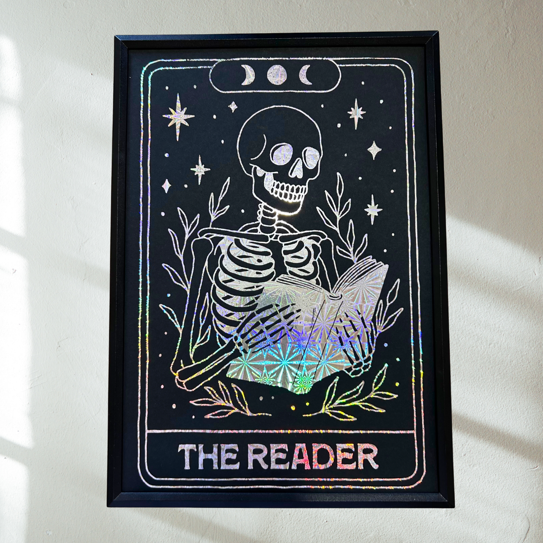 A framed tarot card art print featuring a white skeleton reading a book on a black background with holographic rainbow foil accents, hung on a light wall.