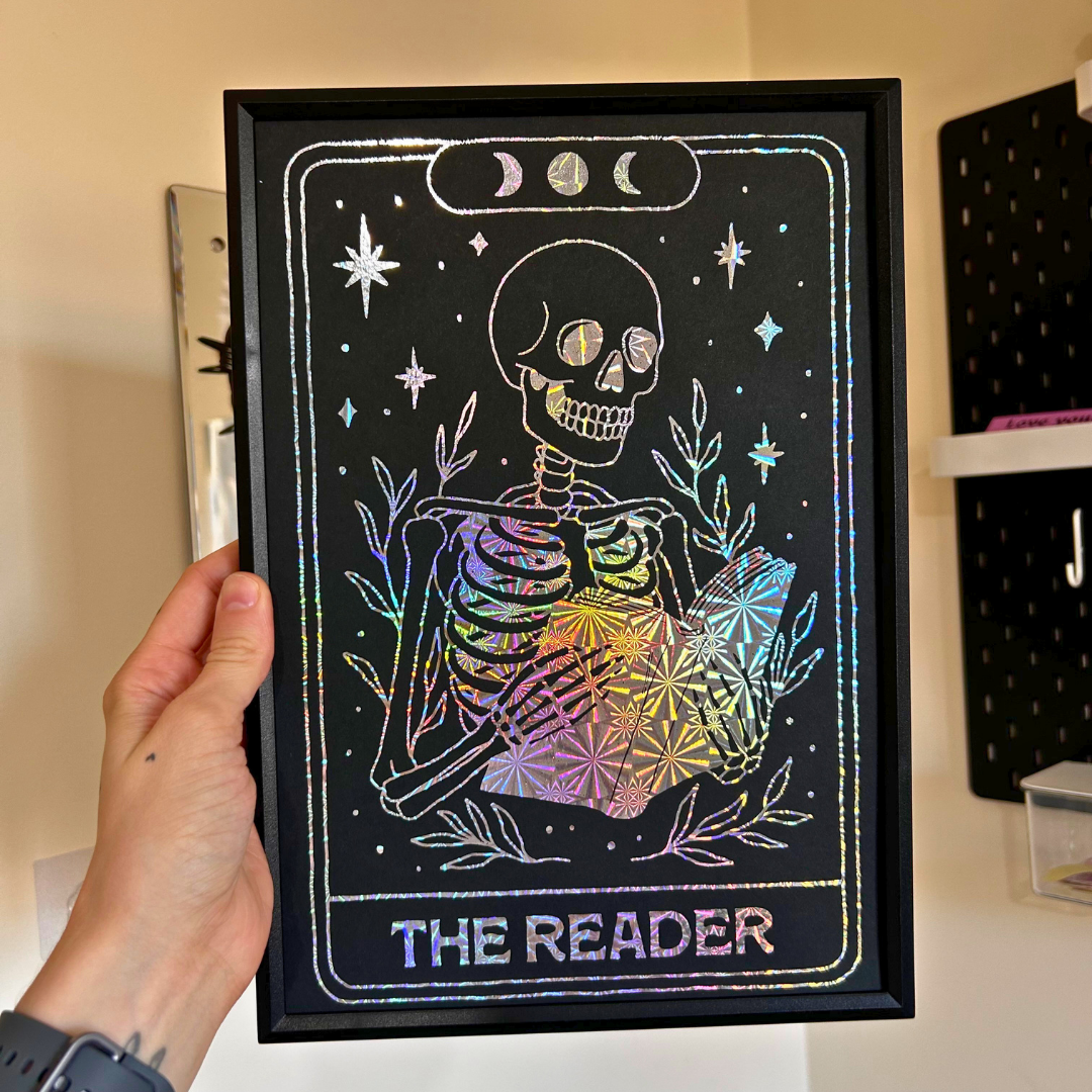 A hand holds a black-framed art print featuring a skeleton card design titled 'THE READER' with holographic rainbow lettering and star details on a black background, shown against an indoor wall shelf setting.