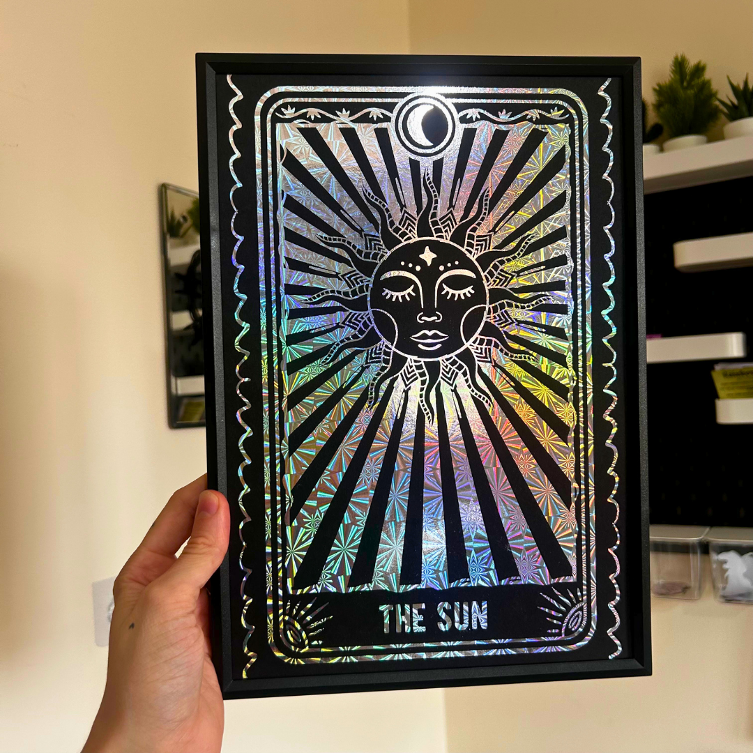 A hand holds a black-framed art print of 'The Sun' tarot card, featuring a holographic silver foil design with a sun face and rays, set against a home interior background.