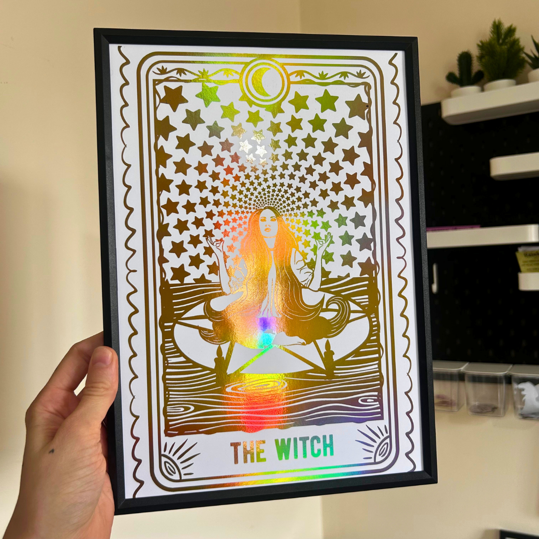 A hand holds a black-framed tarot card art print featuring 'The Witch' with a holographic rainbow foil overlay. The card design shows a meditating witch surrounded by stars and a rainbow spectrum.
