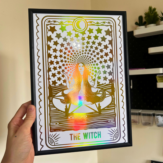 A hand holds a black-framed tarot card art print featuring 'The Witch' with a holographic rainbow foil overlay. The card design shows a meditating witch surrounded by stars and a rainbow spectrum.