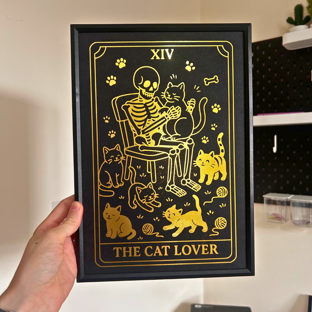 A hand holds a framed art print featuring a tarot card design titled 'THE CAT LOVER'. The print has a black background with gold illustrations of a skeleton sitting on a chair with several cats around it.