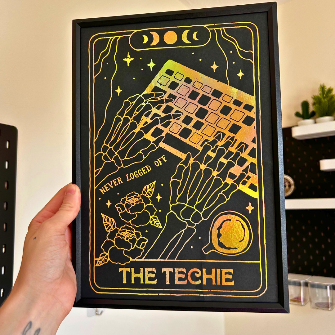 A hand holds a black framed art print featuring two skeleton hands typing on a laptop with gold and iridescent foil details and the text 'THE TECHIE' and 'NEVER LOGGED OFF'.