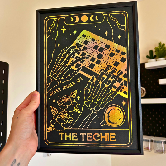 A hand holds a black framed art print featuring two skeleton hands typing on a laptop with gold and iridescent foil details and the text 'THE TECHIE' and 'NEVER LOGGED OFF'.