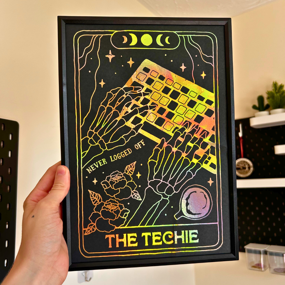 A hand holds a framed art print featuring a stylized card-art design with skeleton hands, a keyboard, moon phases, flowers, and the text 'NEVER LOGGED OFF' and 'THE TECHIE' in a neon gradient on a black background.