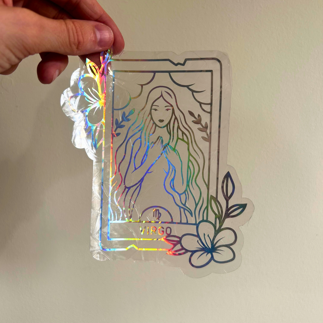 A hand holds a holographic sticker shaped like a tarot card with a line-art illustration of a woman, floral accents, and the word 'Virgo' at the bottom, shown against a plain beige wall.