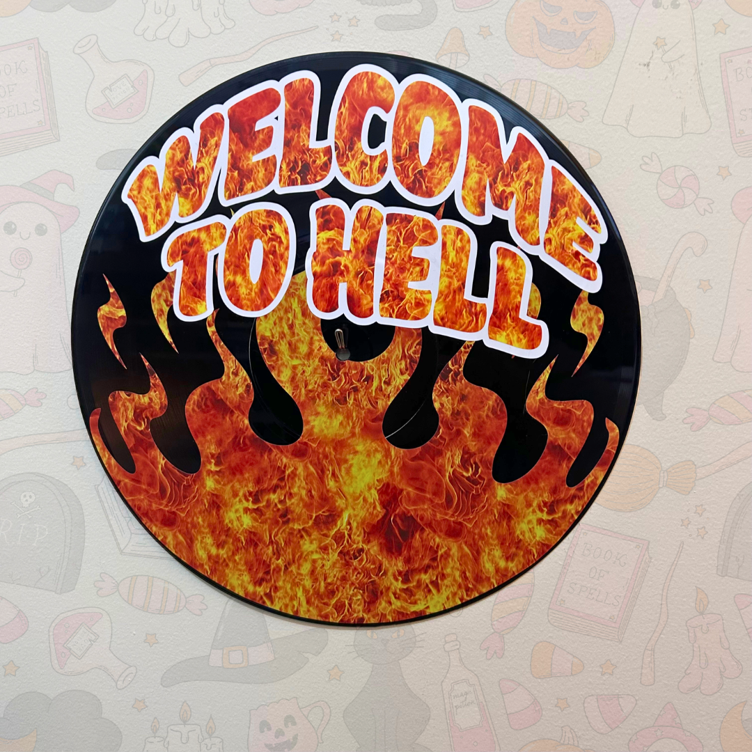 A circular wall sign featuring the phrase 'WELCOME TO HELL' in bold orange and yellow flame-style lettering with white outlines, set against a black background with red and orange flames. The sign is displayed on a light beige wall with faint Halloween-themed illustrations.