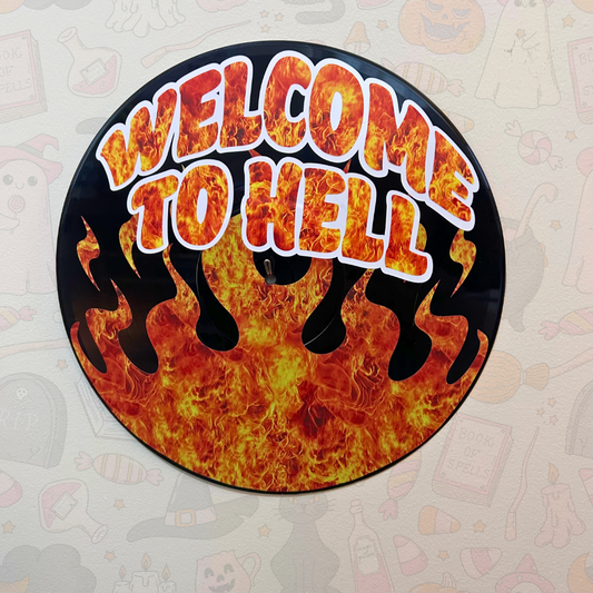 A circular wall sign featuring the phrase 'WELCOME TO HELL' in bold orange and yellow flame-style lettering with white outlines, set against a black background with red and orange flames. The sign is displayed on a light beige wall with faint Halloween-themed illustrations.